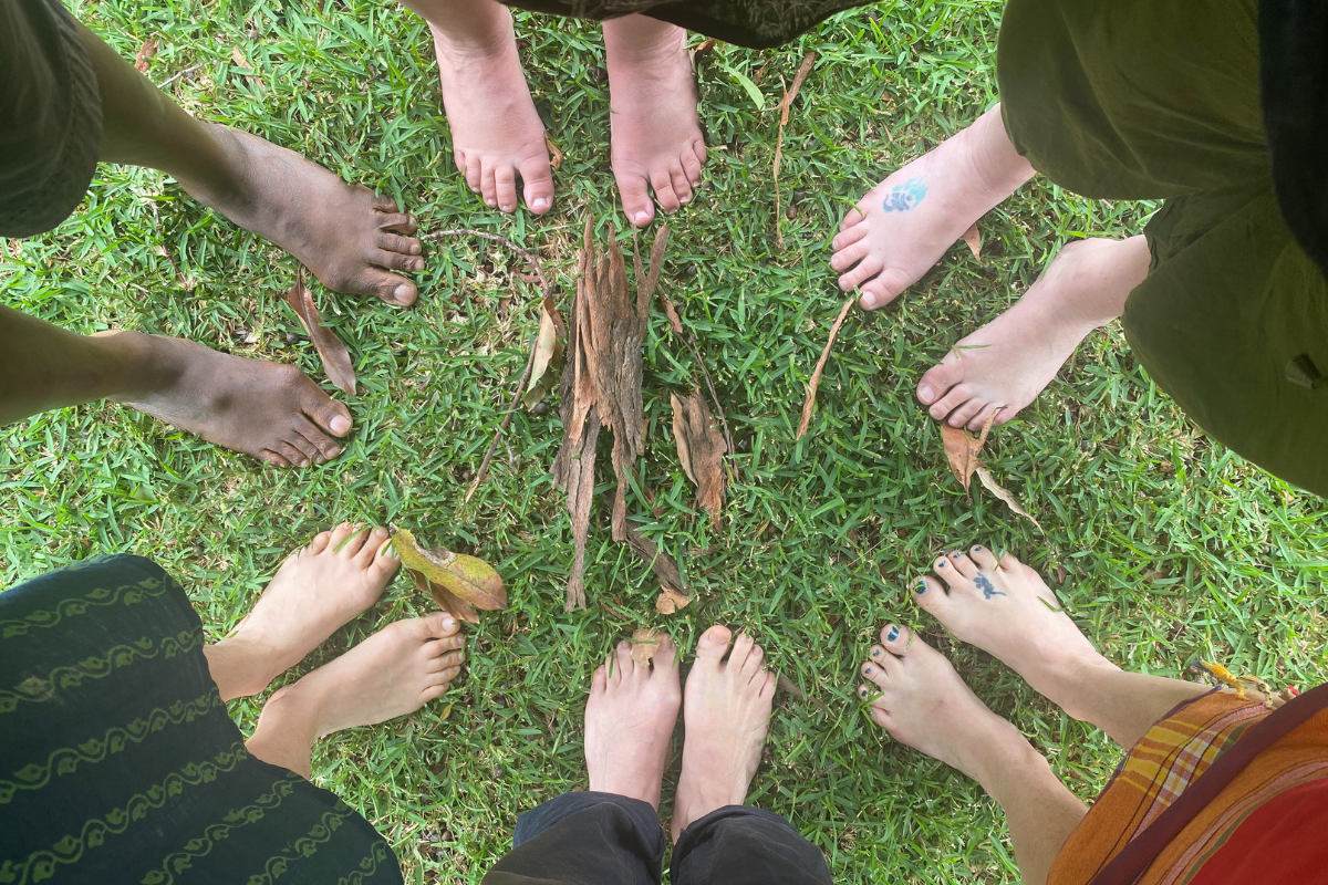 Six pairs of bare feet standing in a circle on green grass, with leaves and bark in the centre.